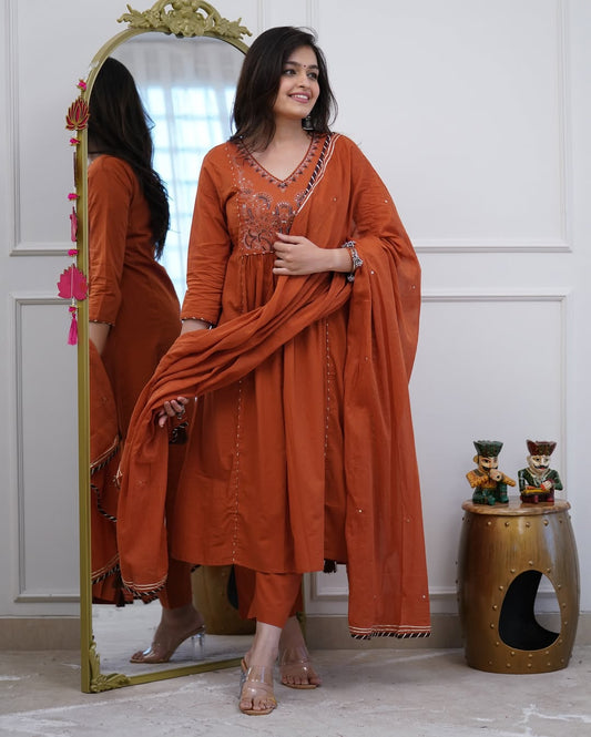 Woman in an orange traditional outfit standing next to a mirror, with a small table and decorative items in the background.